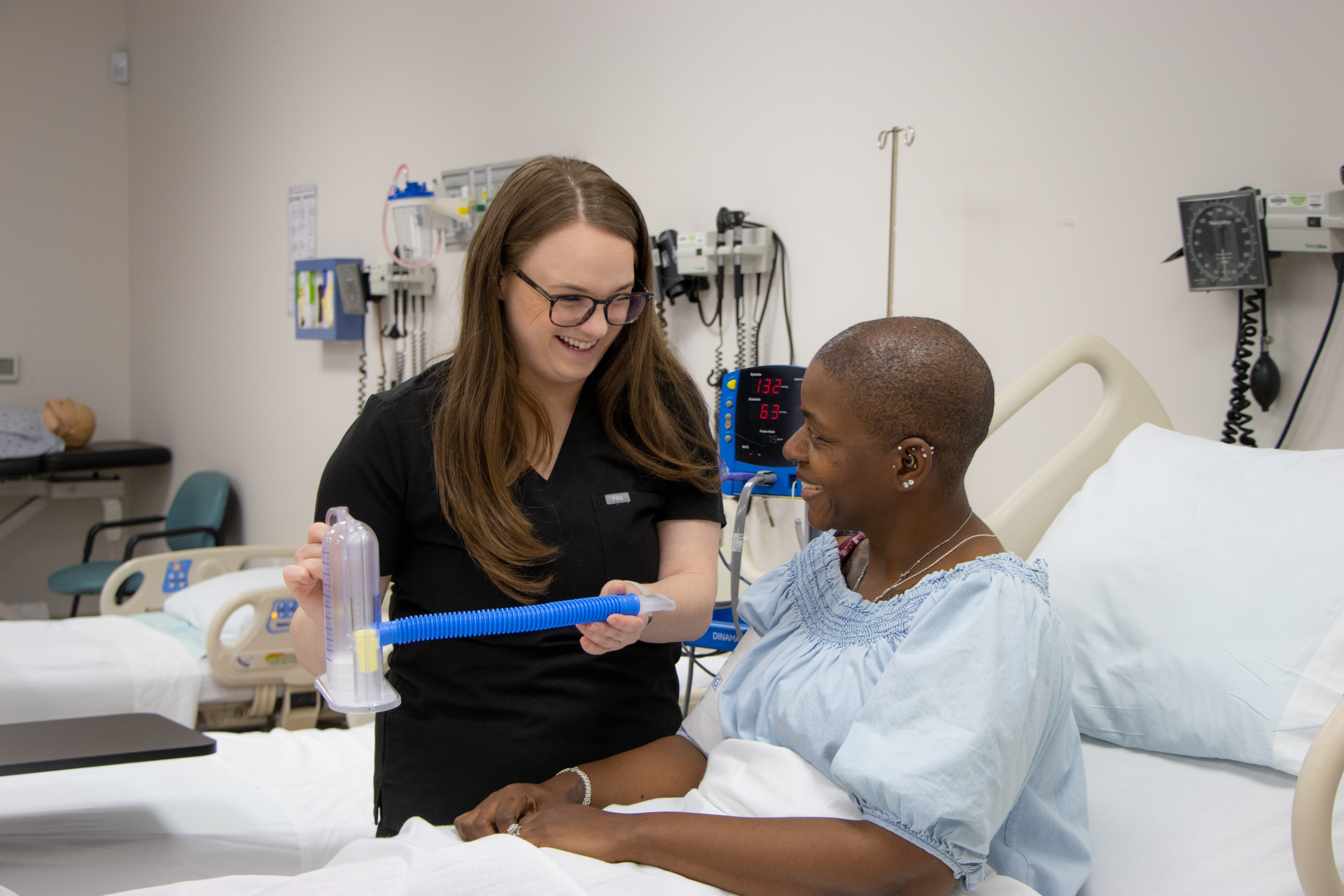 Female Respiratory instructor showing female student, seated in a hospital bed, a tool in a lab.