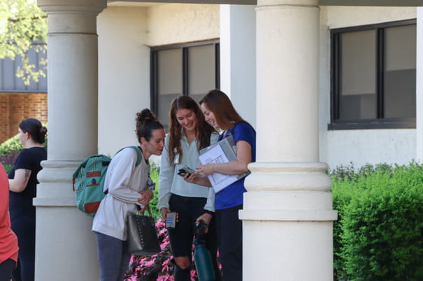 Three female students smiling and looking at a phone screen together under a covered walkway with columns.