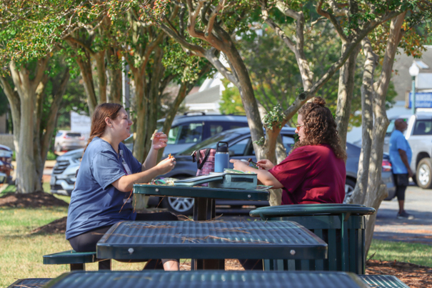 Two female students sitting at a picnic table outside, eating lunch.